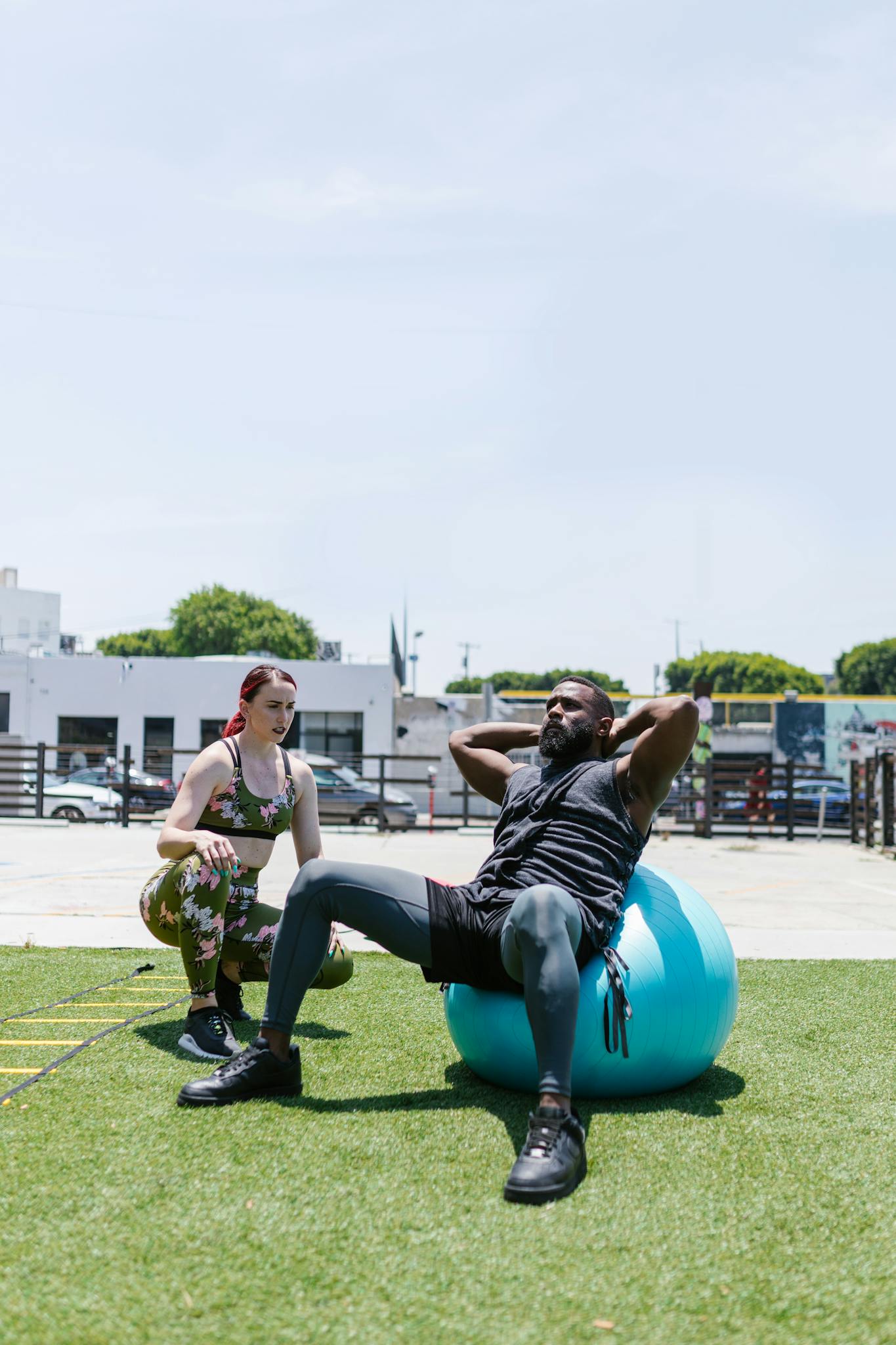 A man doing sit-ups on an exercise ball outdoors with a trainer guiding him, promoting fitness.